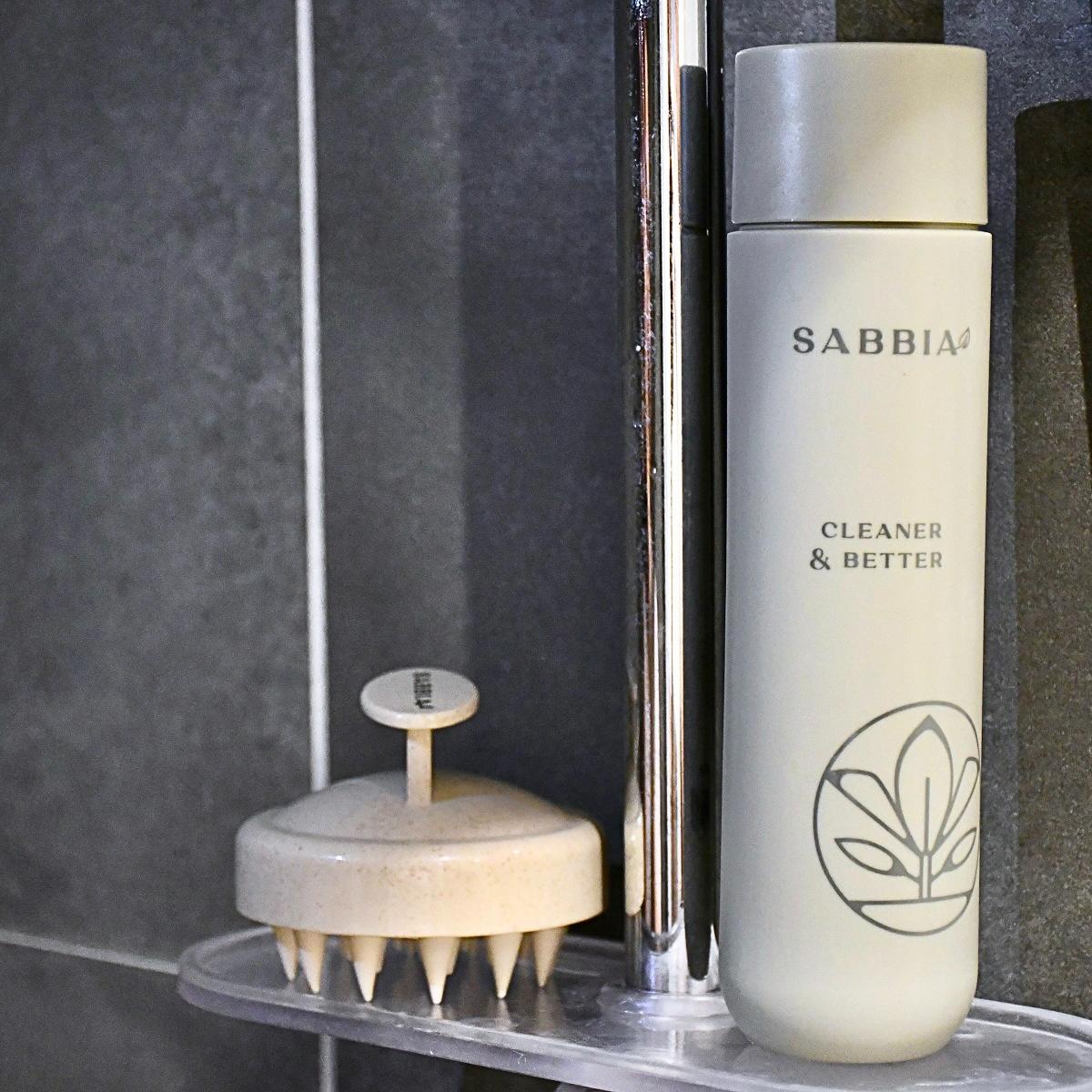 Sabbia šampon a masážní kartáč na vlasy umístěné na sprchové poličce v koupelně.