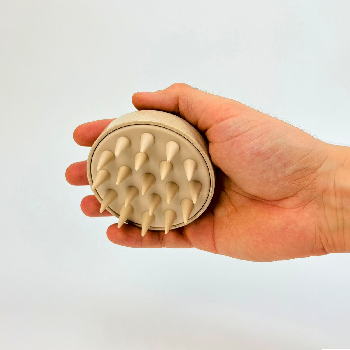 Ruka držiaca béžový silikónový masážny hrebeň na pokožku hlavy s mäkkými hrotmi, na bielom pozadí.