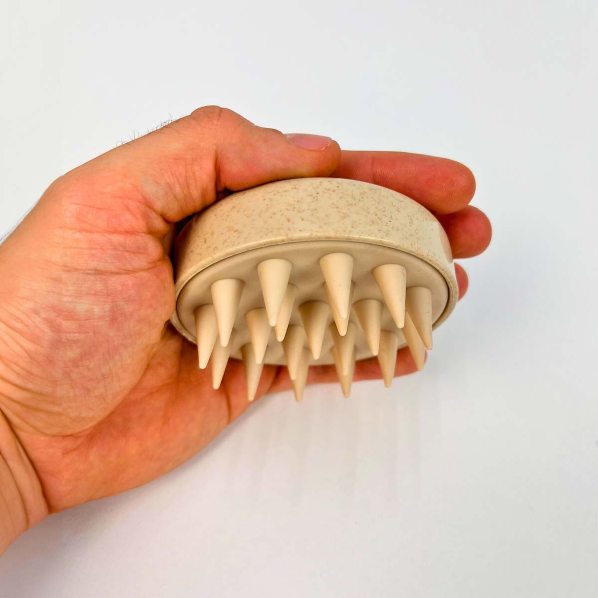 Ruka držící béžový silikonový masážní kartáč na pokožku hlavy s měkkými hroty, na bílém pozadí.